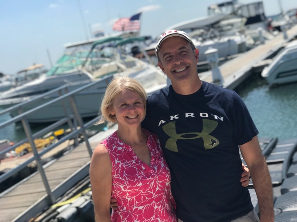 Smiling man and woman at marina with boats and US flag behind.