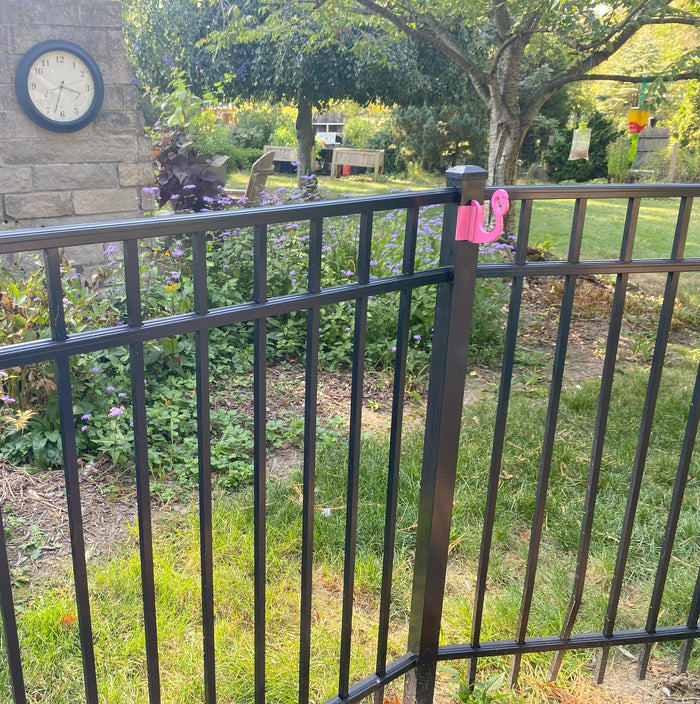 A pink flamingo hook clamped on a black aluminum fence post in a garden with a clock nearby.