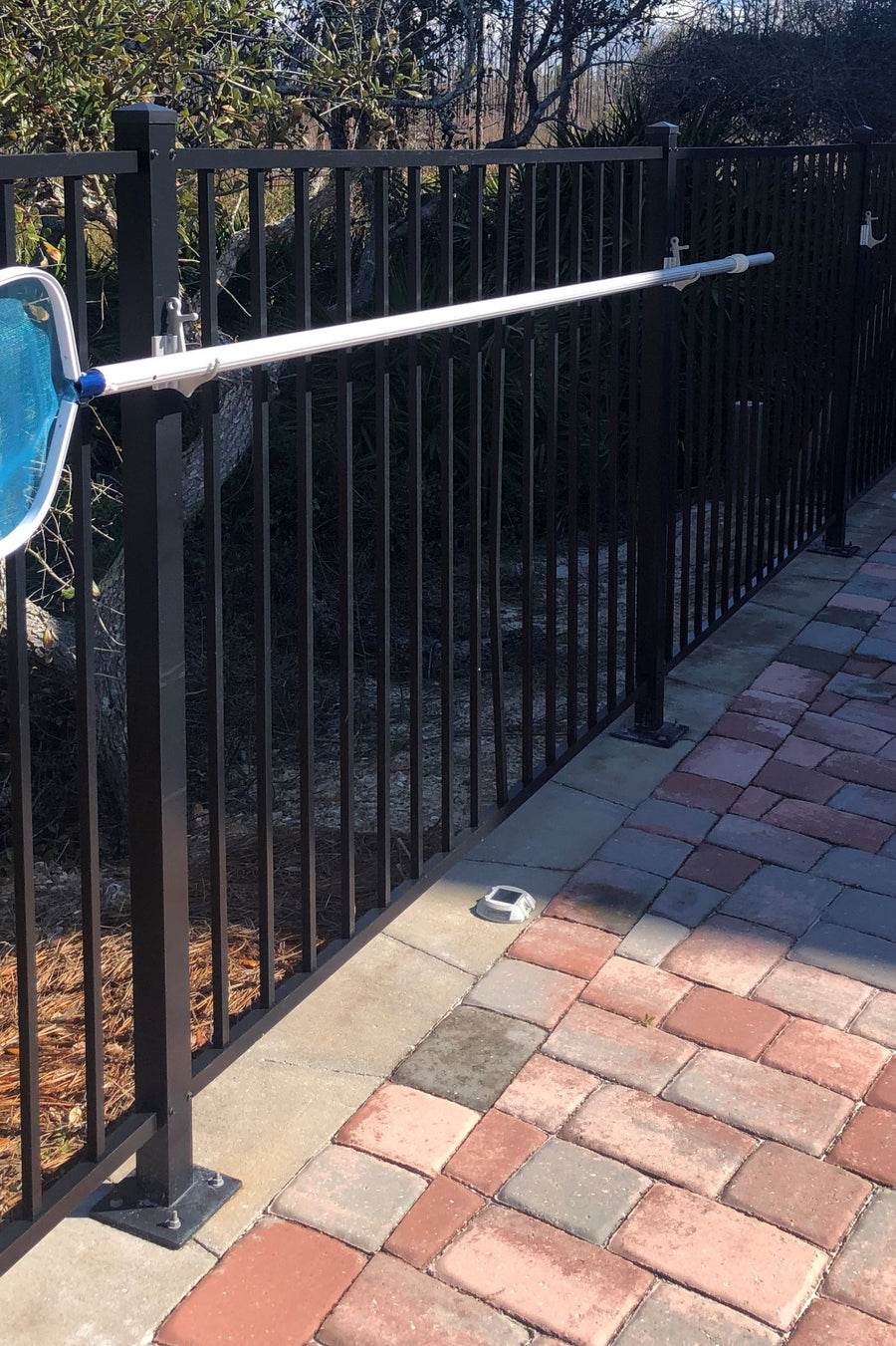 Black fence along brick path, pool pole resting on clamp hooks.
