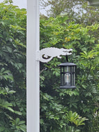 White alligator hanger on a white post holding a black lantern against green leafy shrubs.