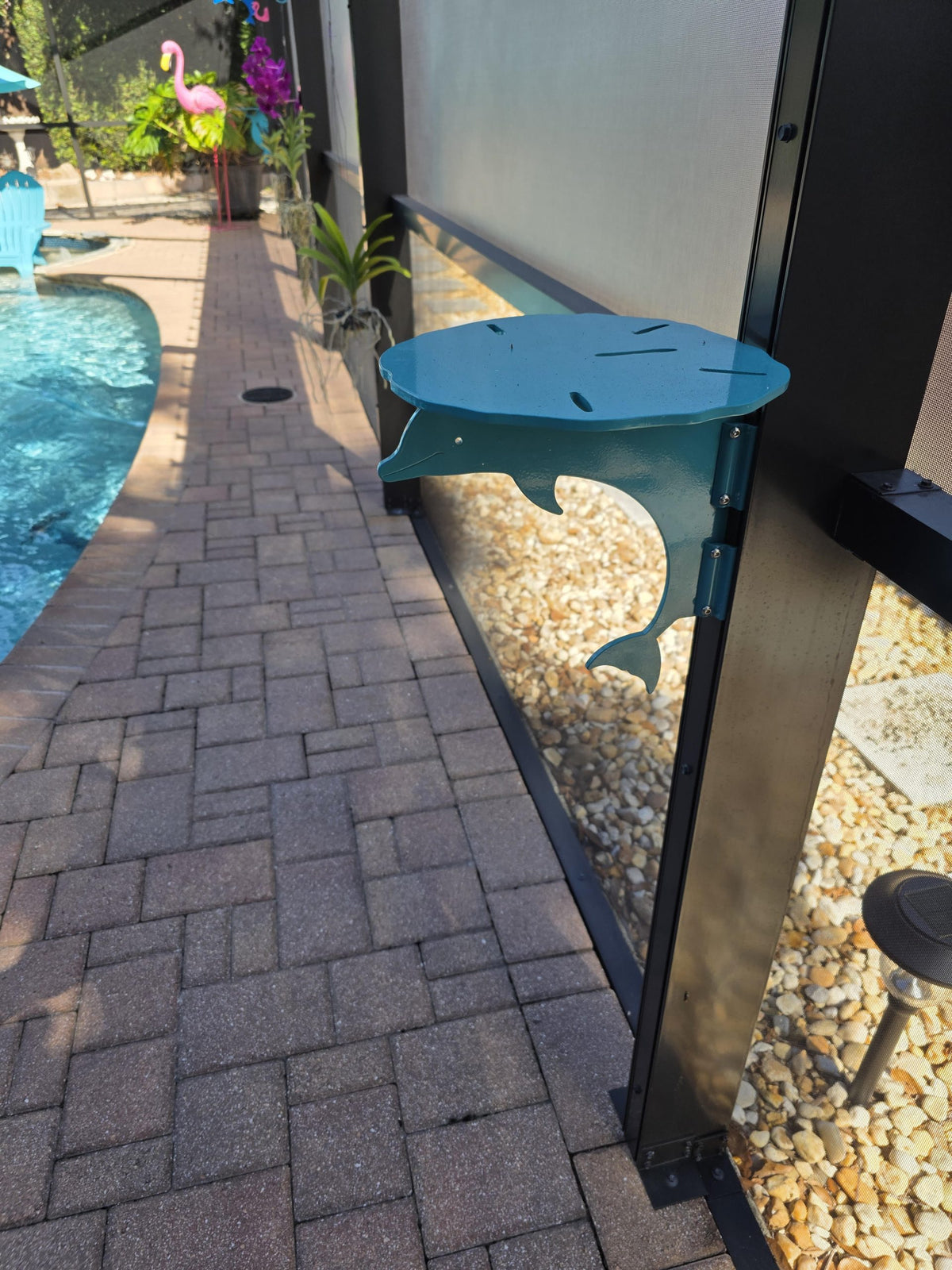 Turquoise dolphin mini table clamped to a dark pool-cage post beside a brick paver pool deck.