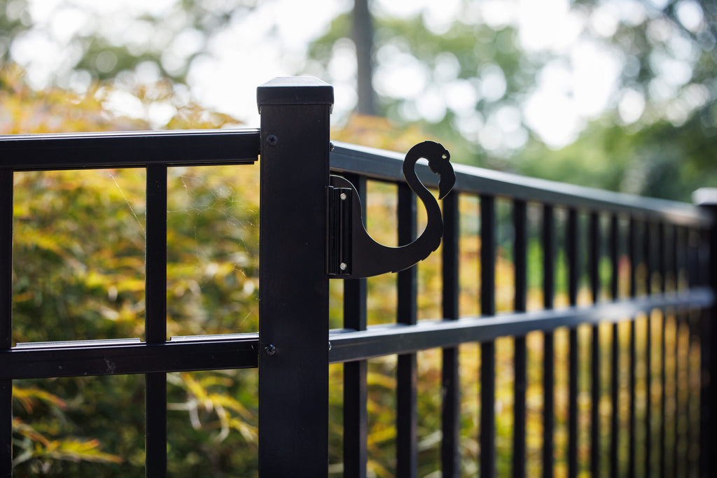 Black flamingo hook clamped to a black aluminum fence post with golden foliage behind.