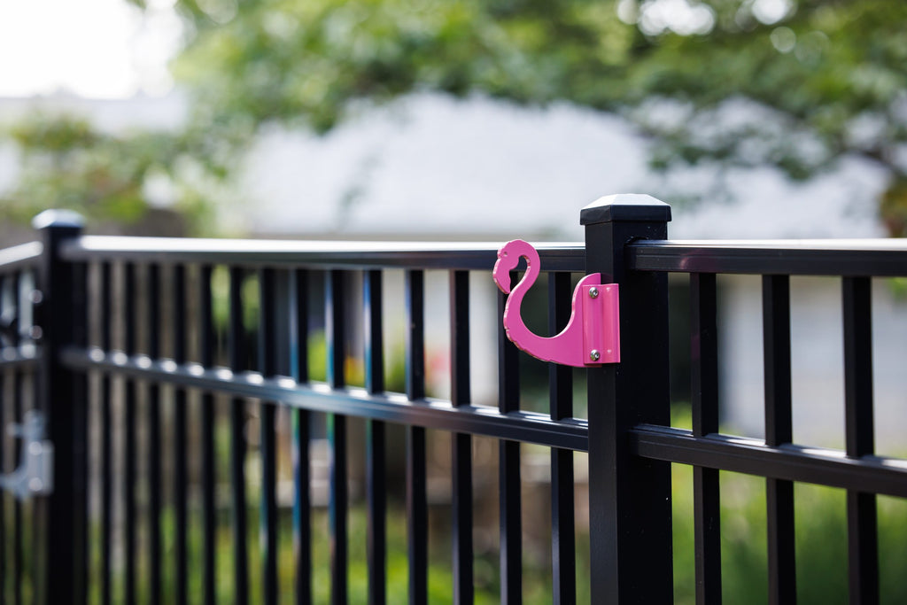 Pink flamingo hook clamped to a black aluminum fence post in a backyard.