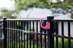 Pink flamingo hook clamped to a black aluminum fence post in a backyard.