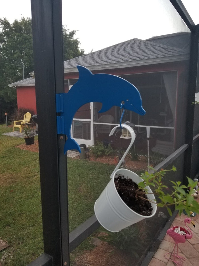 A blue dolphin hanger on a dark lanai post holding a white plant pot with a house beyond.
