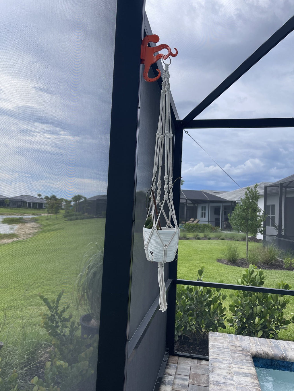 Orange octopus hook on a dark lanai post holding a white macrame plant hanger, lawn beyond.