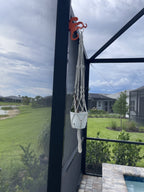 Orange octopus hook on a dark lanai post holding a white macrame plant hanger, lawn beyond.