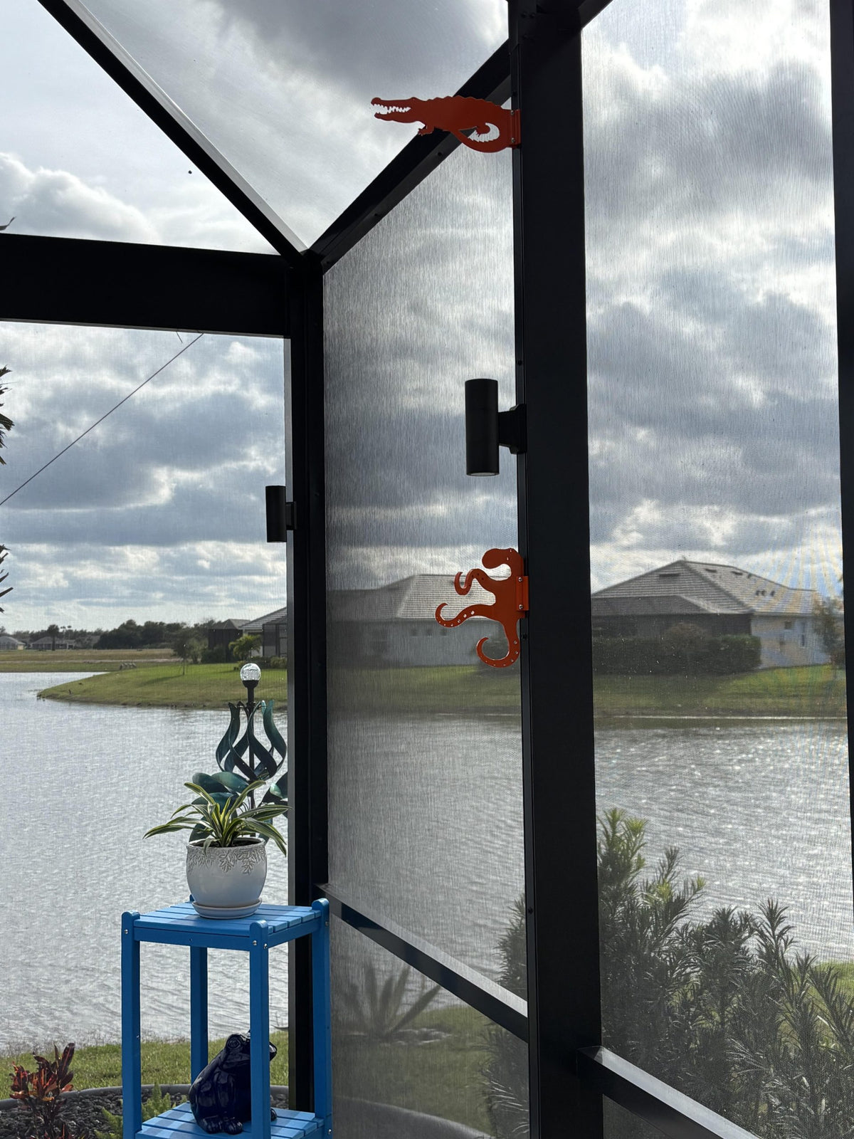 Two orange octopus hooks on dark screen-enclosure posts overlooking a lake, a blue plant stand below.