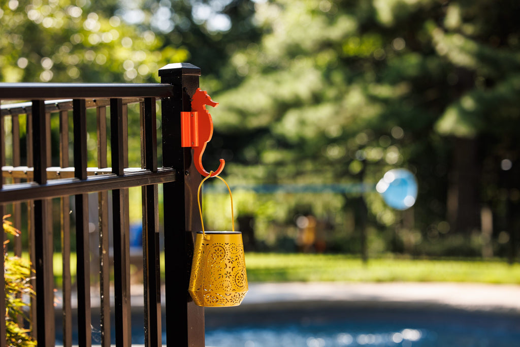 Orange seahorse hook on a black aluminum fence post holding a yellow lantern near a pool.