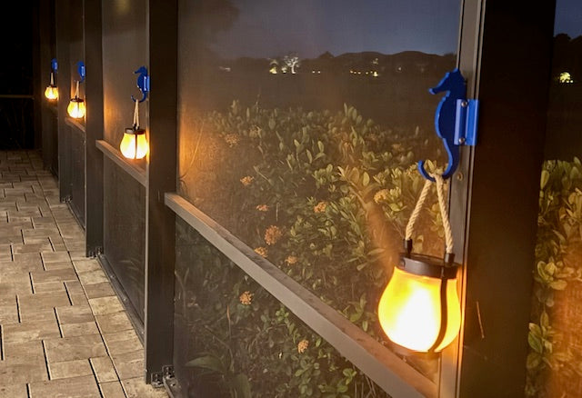 Blue seahorse hooks on dark lanai posts holding glowing lanterns along a planter ledge at dusk.