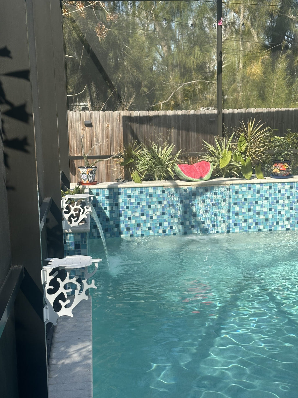 Two white turtle-shaped mini tables clamped to dark lanai posts beside a tiled pool wall.