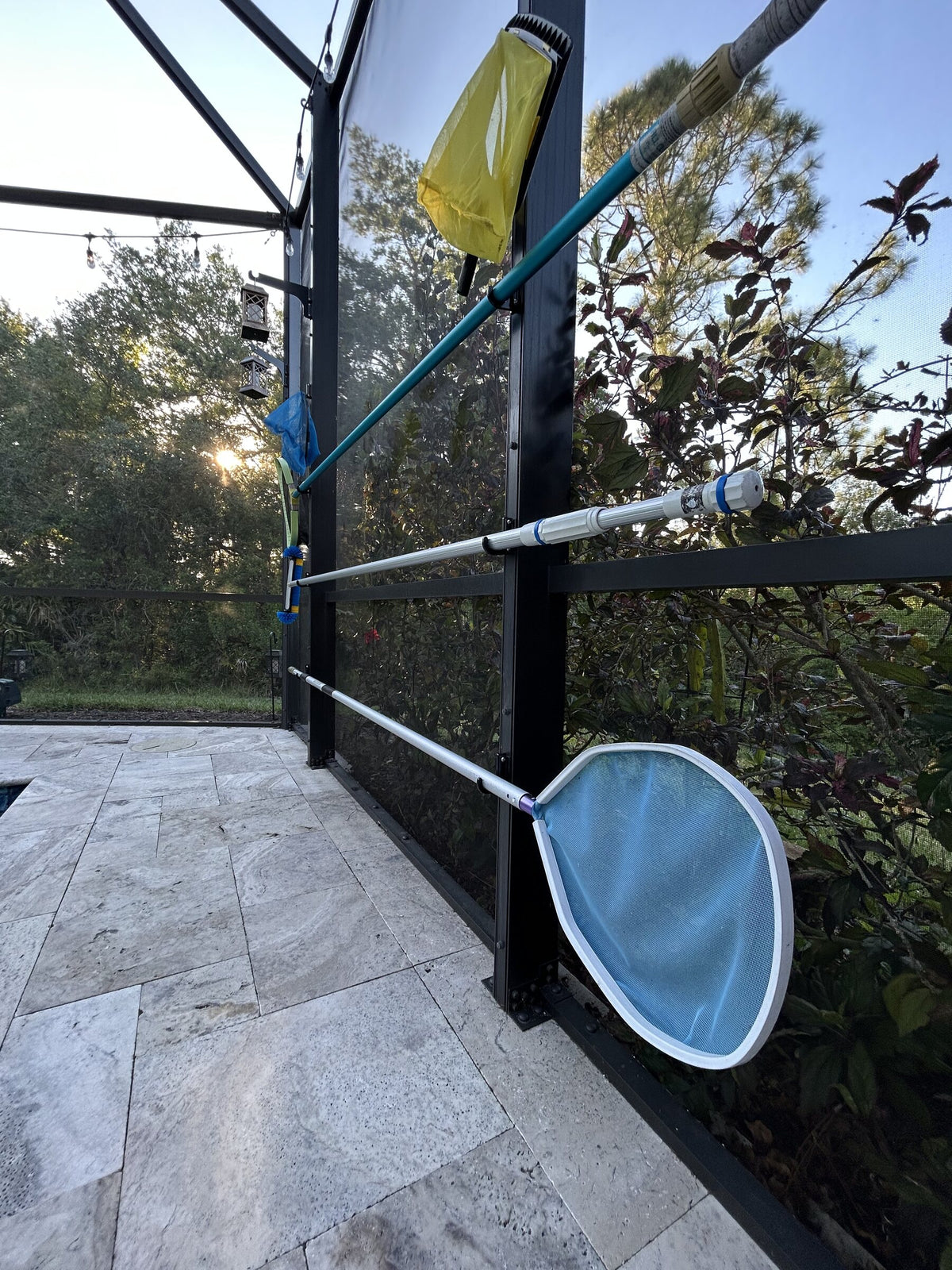 A pool skimmer net, brush, and poles resting on clamp-on hooks along dark screen-enclosure posts.