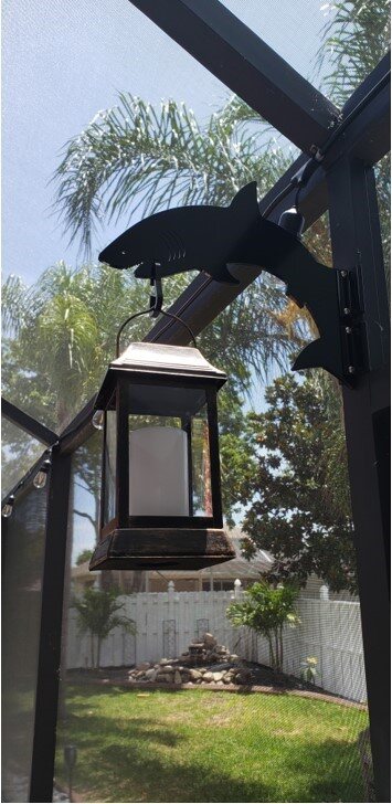 Black shark-shaped hanger on a dark pool-cage post holding a lantern with palm trees behind.